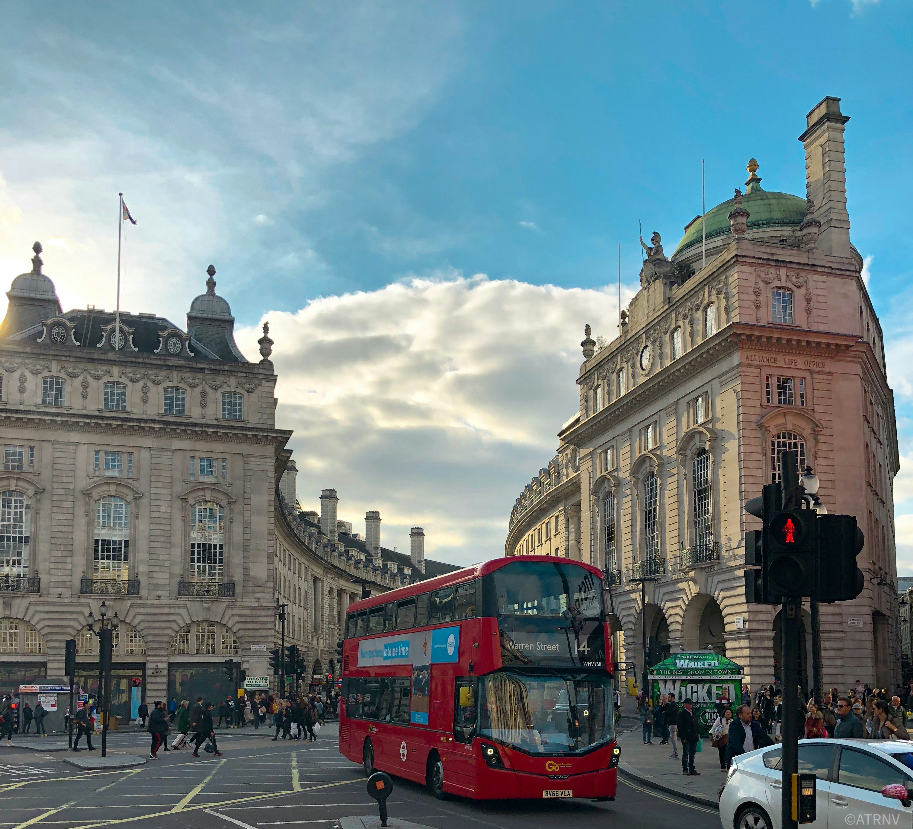 Piccadilly Circus, London — a red double-decker bus crossing the junction beneath the Alliance Life Office building at golden hour.
